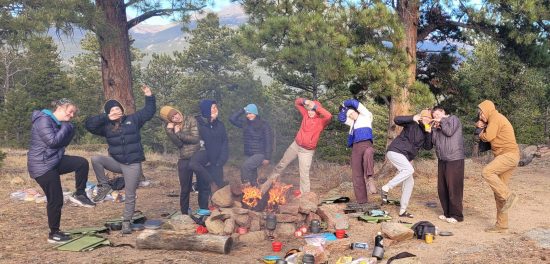 Cottonwood Alumni stand around the campfire in unusual positions.