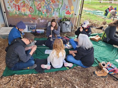 Attendees of the Ancestral Skills Gathering sit on the ground in a circle while learning to make beads.