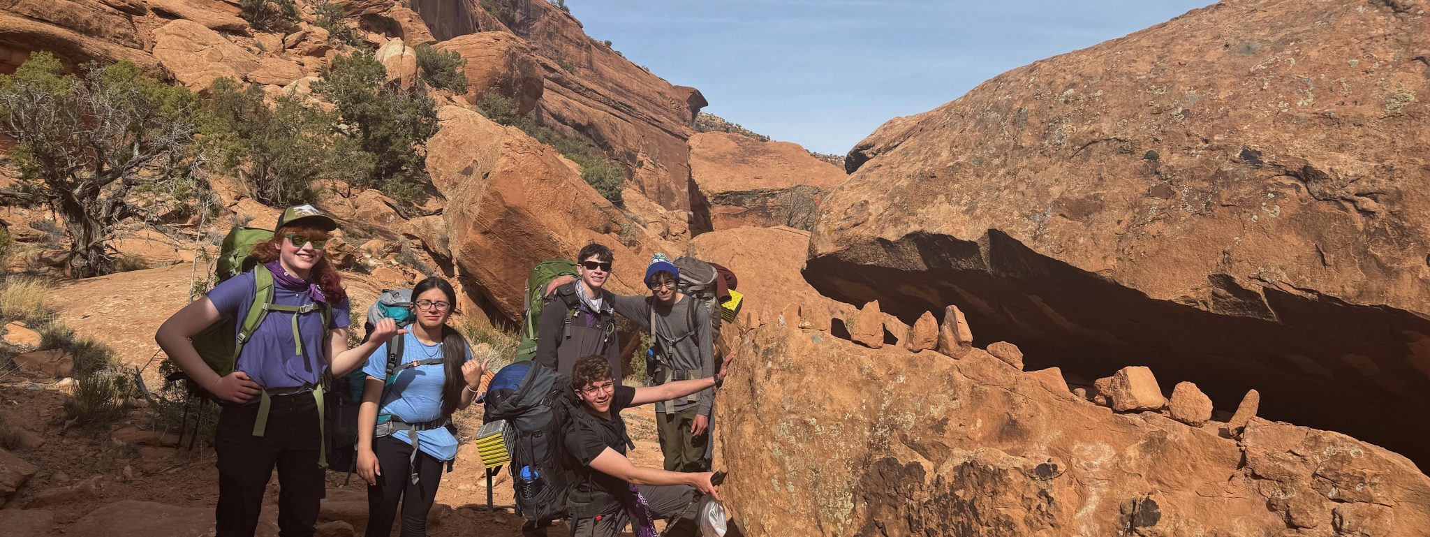 During their canyon backpacking trip, tudents pose near a huge rock that looks like a creature with teeth.