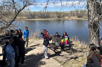 Students swing at a Piñata on the shore of a pond.
