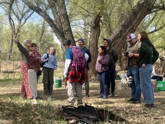 Skills Gathering participants gather around an Instructor among the trees along the creek, building outdoor skills and community connections.