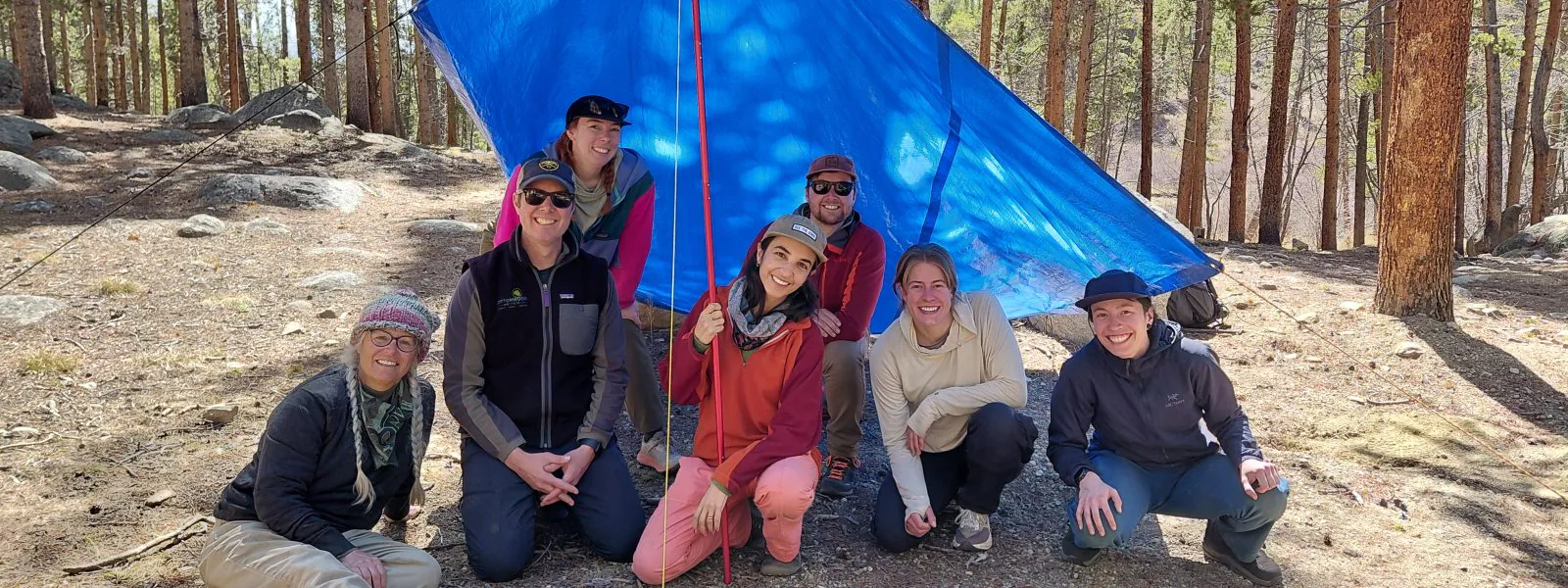 Cottonwood staff pose underneath their tarp shelter after learning new techniques for building shelters during spring training.