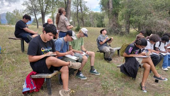 Students sit on benches with clipboards as they learn about hydrology.