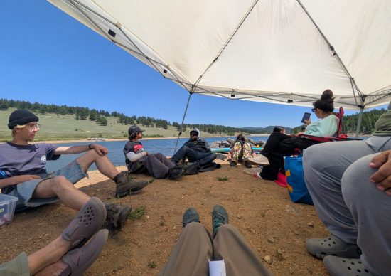 Casa family camping participants sit in a circle under a shade tent near a reservoir.