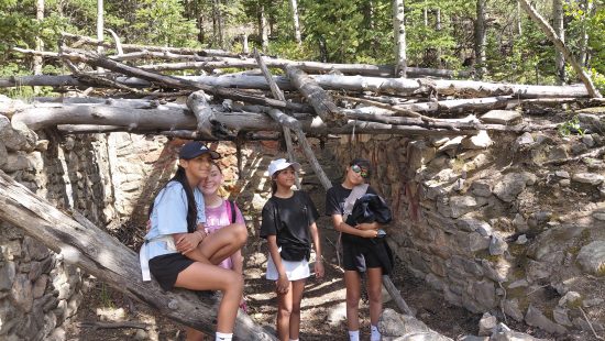 Four LYC students pose under a shelter built from logs and sticks.