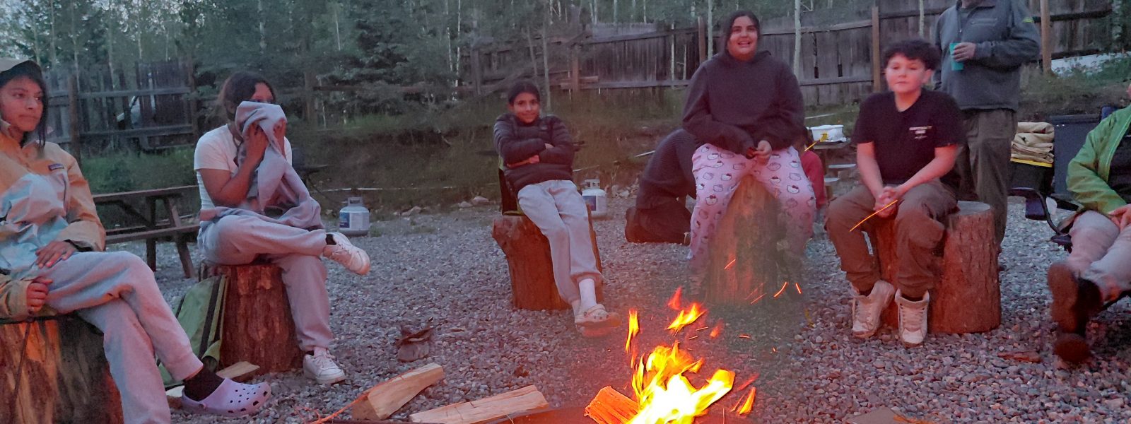 Longmont Youth Center group sits around a campfire at dusk.