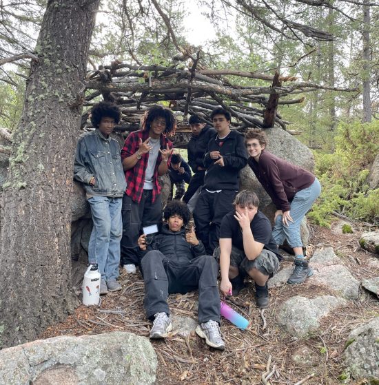 Students pose under their shelter built from logs found outdoors.